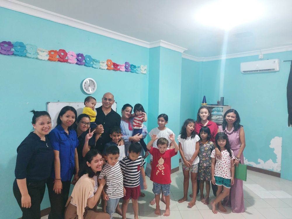 Group of children and adults smiling in front of a light blue wall.